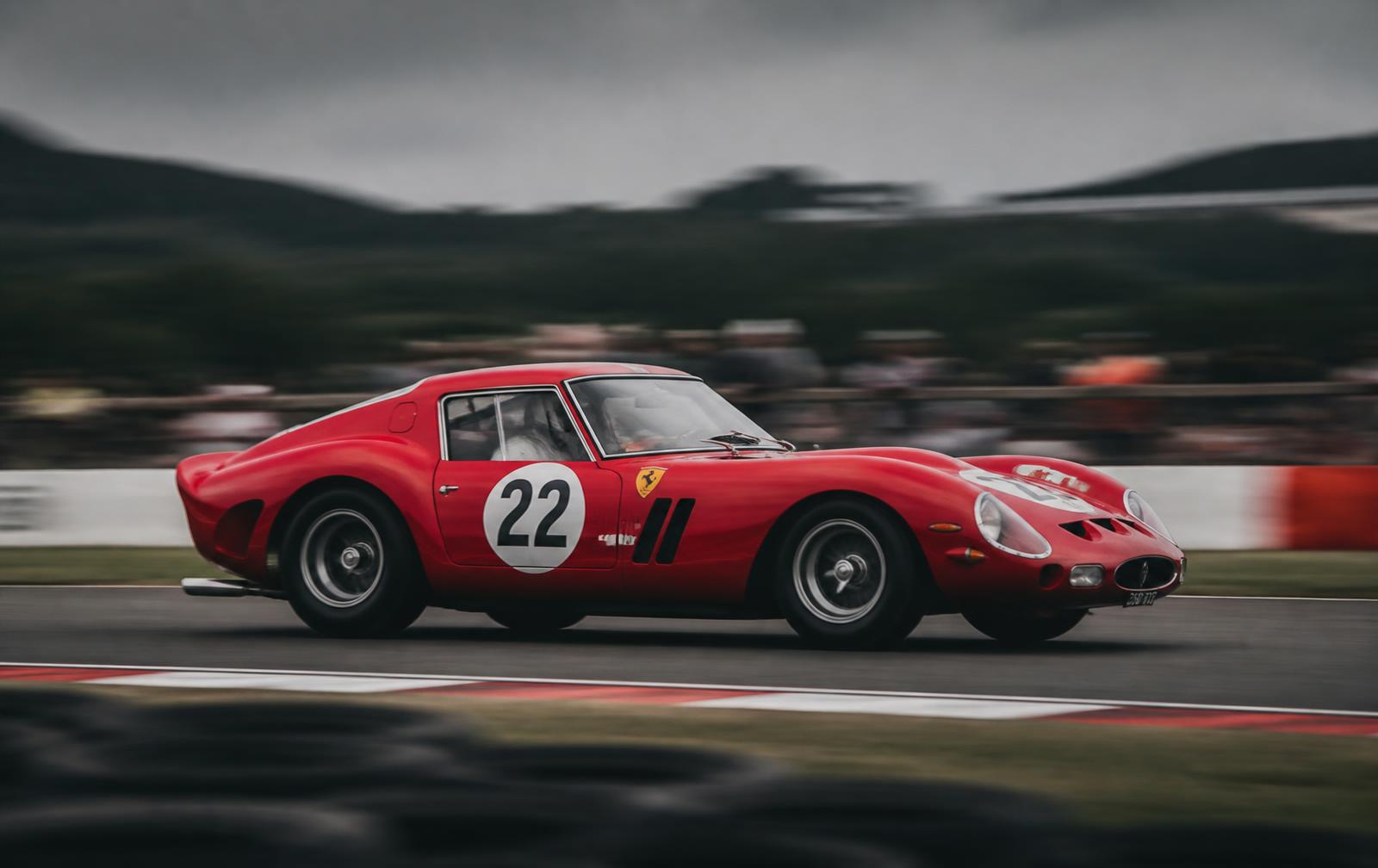 Red 1960s racing car panning at speed on a British circuit under overcast light